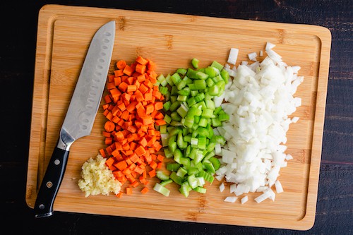 Overhead View of Chopped Vegetables on a Bamboo Cutting Board: Finely chopped carrots, celery, onions, and garlic on a wooden cutting board with a santoku knife